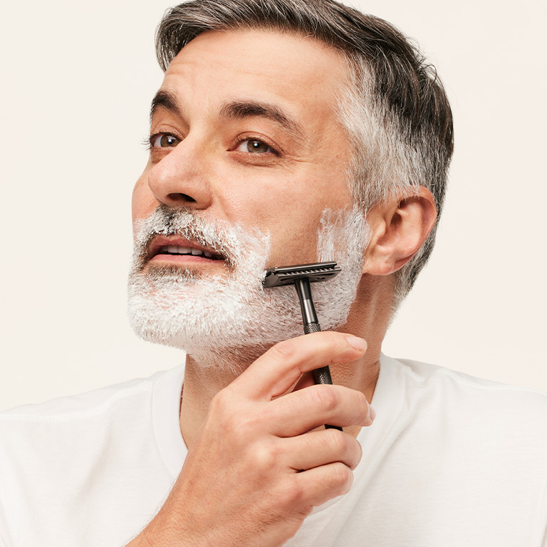 Person wearing a plain white crew-neck shirt holding a razor near the chin against a neutral background